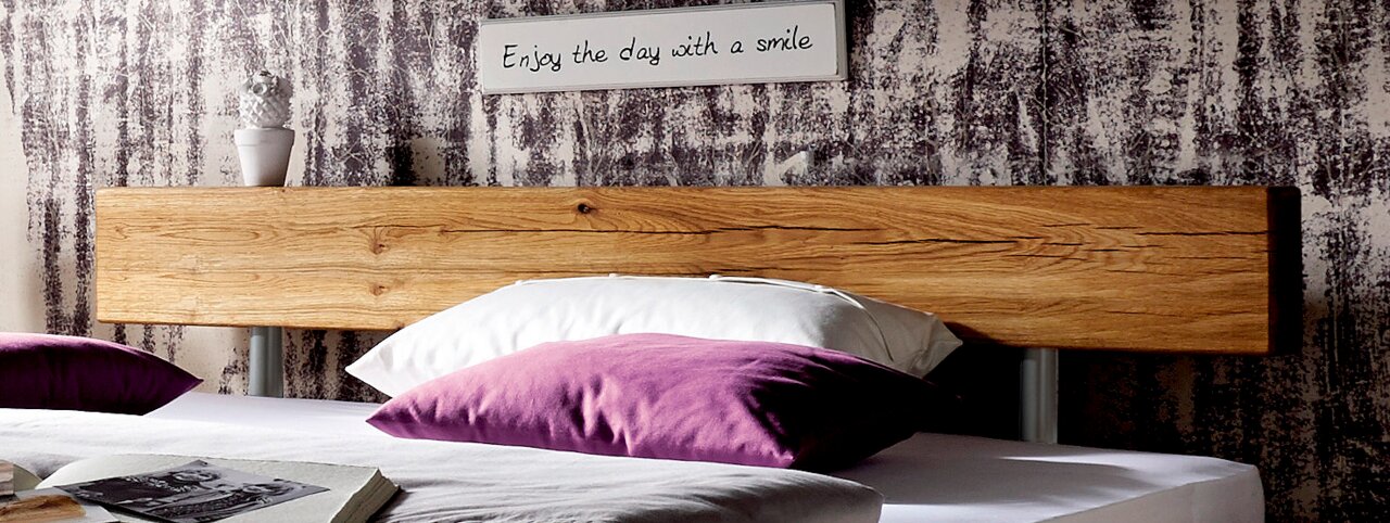 Close-up van een houten bed met een rustiek houten hoofdeinde, versierd met paarse en witte kussens, op de voorgrond. Op de achtergrond een bord met de tekst 'Enjoy the day with a smile'.