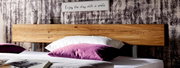 Close-up van een houten bed met een rustiek houten hoofdeinde, versierd met paarse en witte kussens, op de voorgrond. Op de achtergrond een bord met de tekst 'Enjoy the day with a smile'.