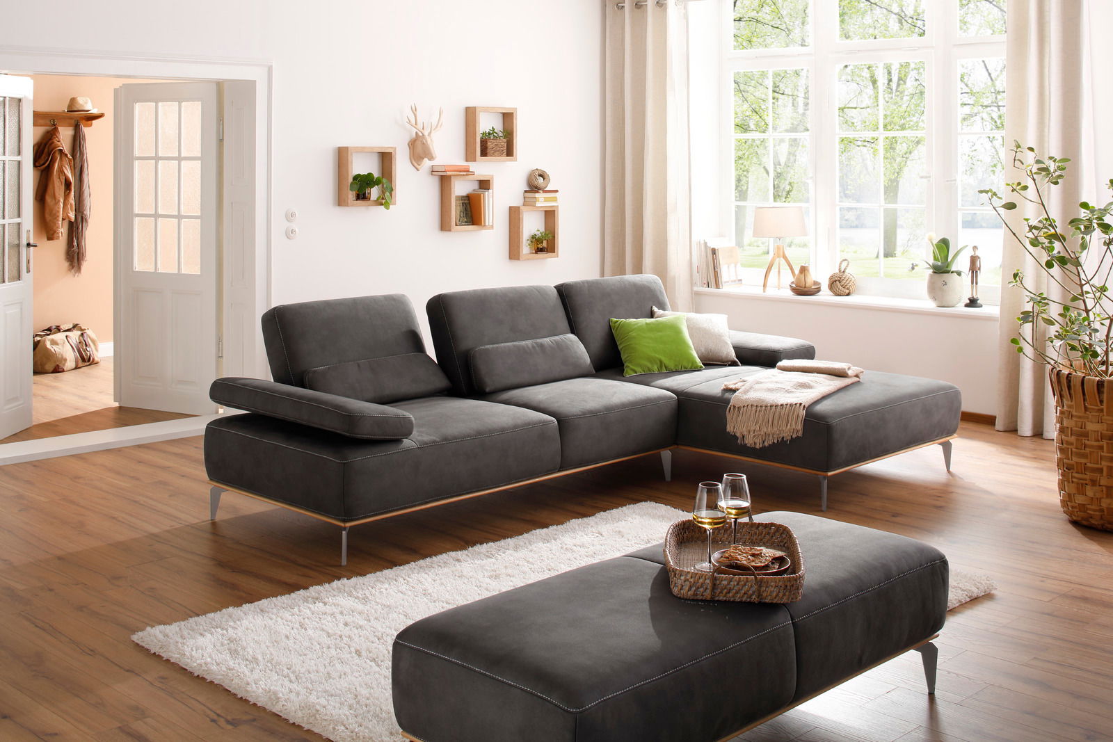 Großer, grauer Hocker in einem modernen Wohnzimmer mit Holzfußboden, neben einem großen Fenster mit Blick ins Grüne, aus seitlicher Perspektive.