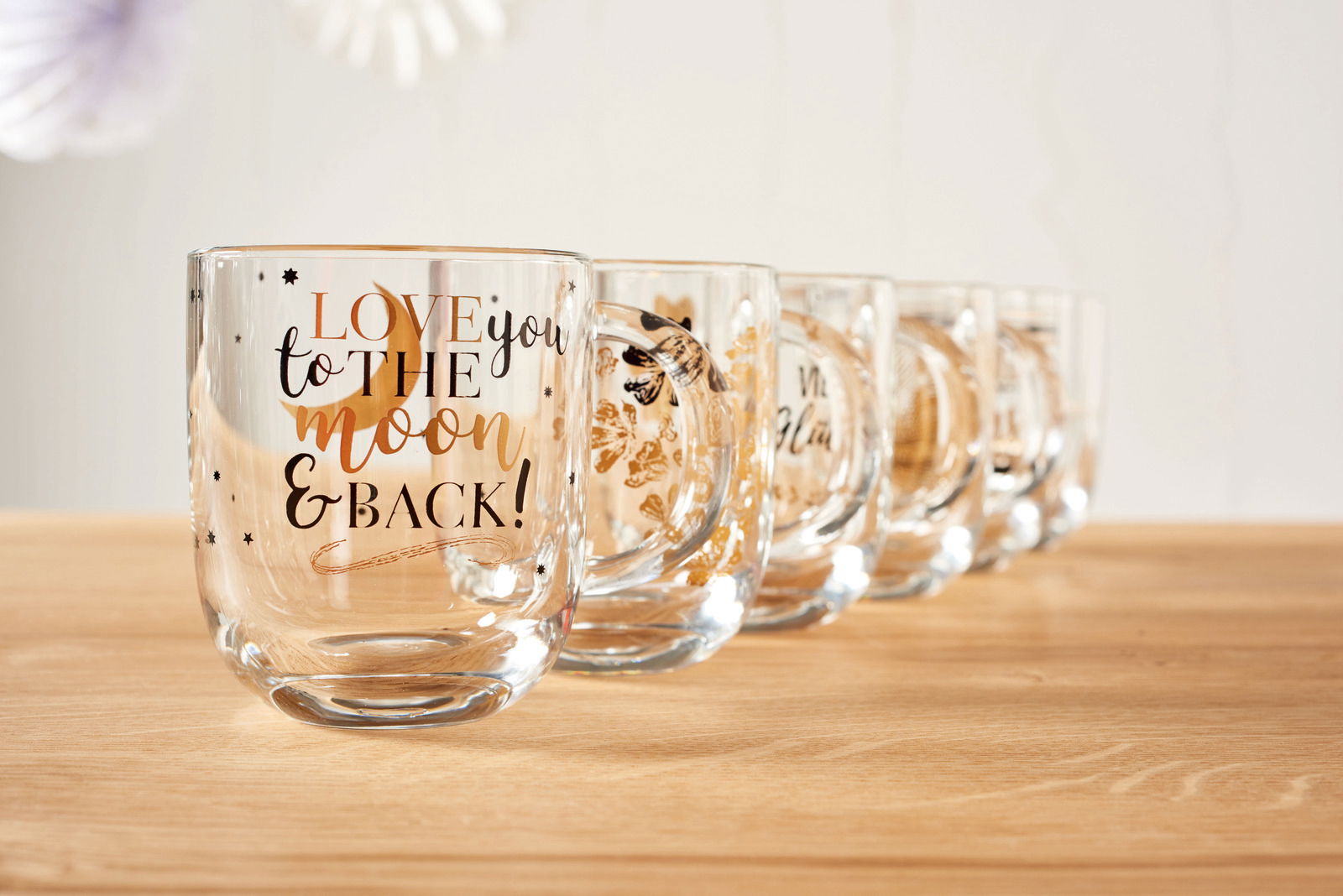 Eine Reihe von durchsichtigen Tassen mit goldener und schwarzer Schrift, die vorderste Tasse zeigt den Text 'LOVE you to the moon & BACK!' in einer schrägen Perspektive.