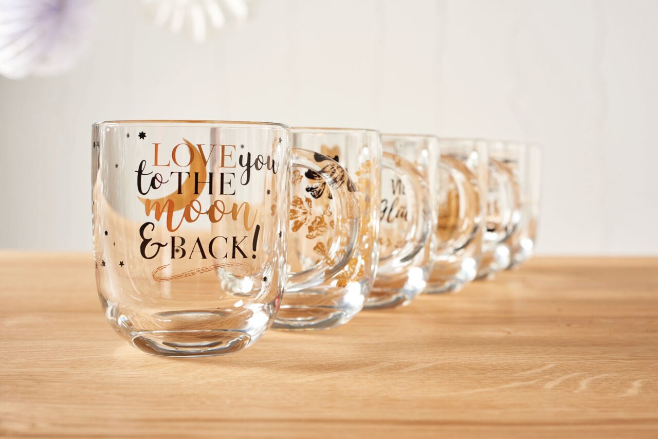 Eine Reihe von durchsichtigen Tassen mit goldener und schwarzer Schrift, die vorderste Tasse zeigt den Text 'LOVE you to the moon & BACK!' in einer schrägen Perspektive.