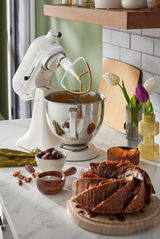 Weiße Küchenmaschine mit Rührschüssel und Rührbesen, seitliche Perspektive, umgeben von Zutaten und einem Kuchen auf der Arbeitsplatte.