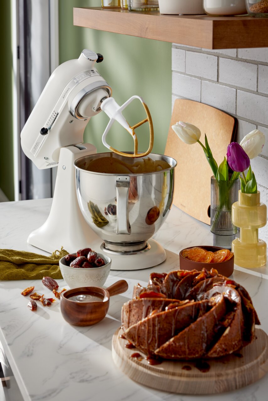 Weiße Küchenmaschine mit Rührschüssel und Rührbesen, seitliche Perspektive, umgeben von Zutaten und einem Kuchen auf der Arbeitsplatte.