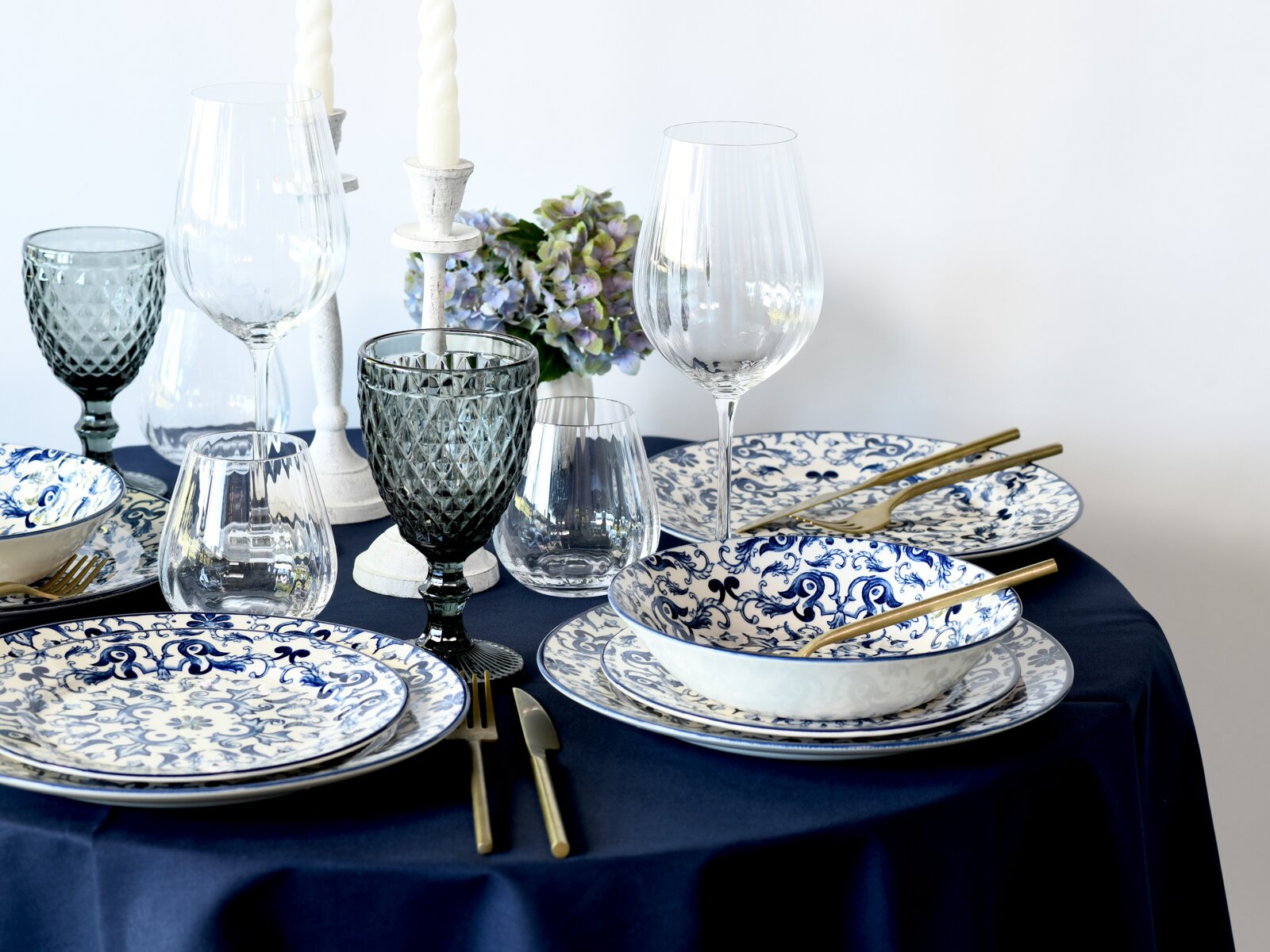 Elegant gedeckter Esstisch mit blau-weiß gemustertem Porzellan, goldfarbenem Besteck, Glaswaren und Kerzenständern auf dunkelblauer Tischdecke, schräg von vorne aus leicht erhöhter Perspektive fotografiert