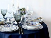 Elegant gedeckter Esstisch mit blau-weiß gemustertem Porzellan, goldfarbenem Besteck, Glaswaren und Kerzenständern auf dunkelblauer Tischdecke, schräg von vorne aus leicht erhöhter Perspektive fotografiert