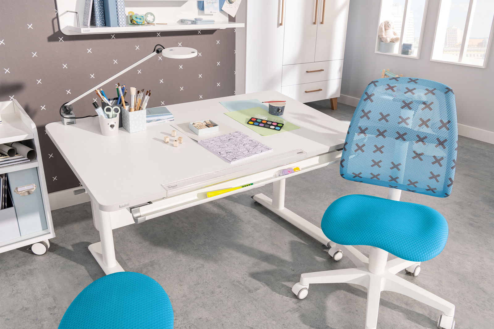 Moderner Jugenddrehstuhl in Blau mit Kreuzmuster, seitliche Perspektive, vor einem weißen Schreibtisch in einem hellen Raum.