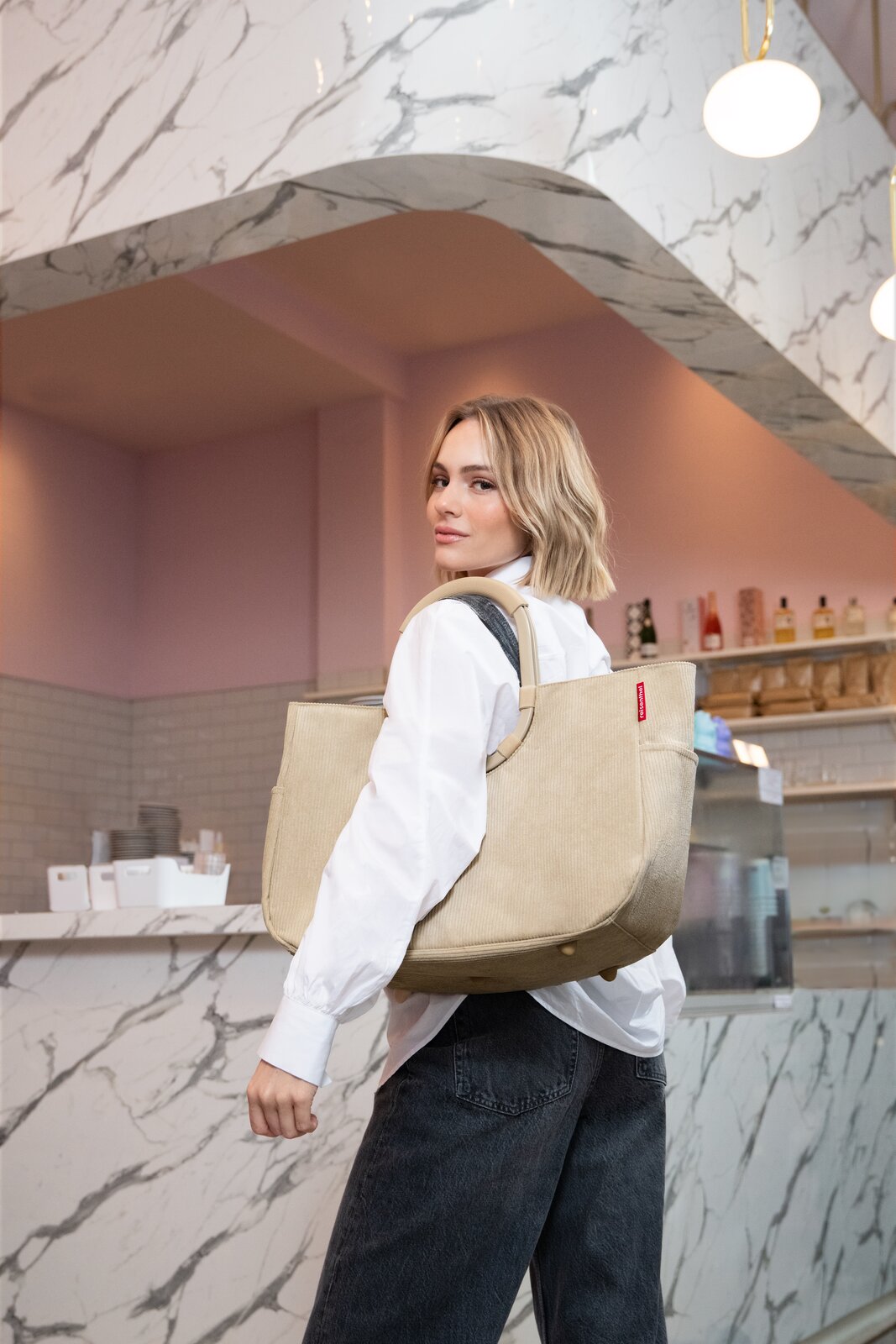 Junge Frau in Café-Umgebung trägt eine große beige Cord-Tote-Bag mit breiten Tragegriffen über der Schulter; Perspektive: leicht seitlich von hinten, Fokus auf die Tasche.