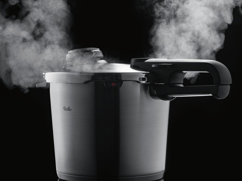 Ein schlanker Schnellkochtopf aus Edelstahl gibt Dampf vor einem dunklen Hintergrund ab.