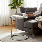 Runder Beistelltisch aus verchromtem Metall neben grauem Sofa, mit Buch, Brille und dampfender Tasse; Perspektive: seitliche Ansicht auf Tisch und Sofaecke.