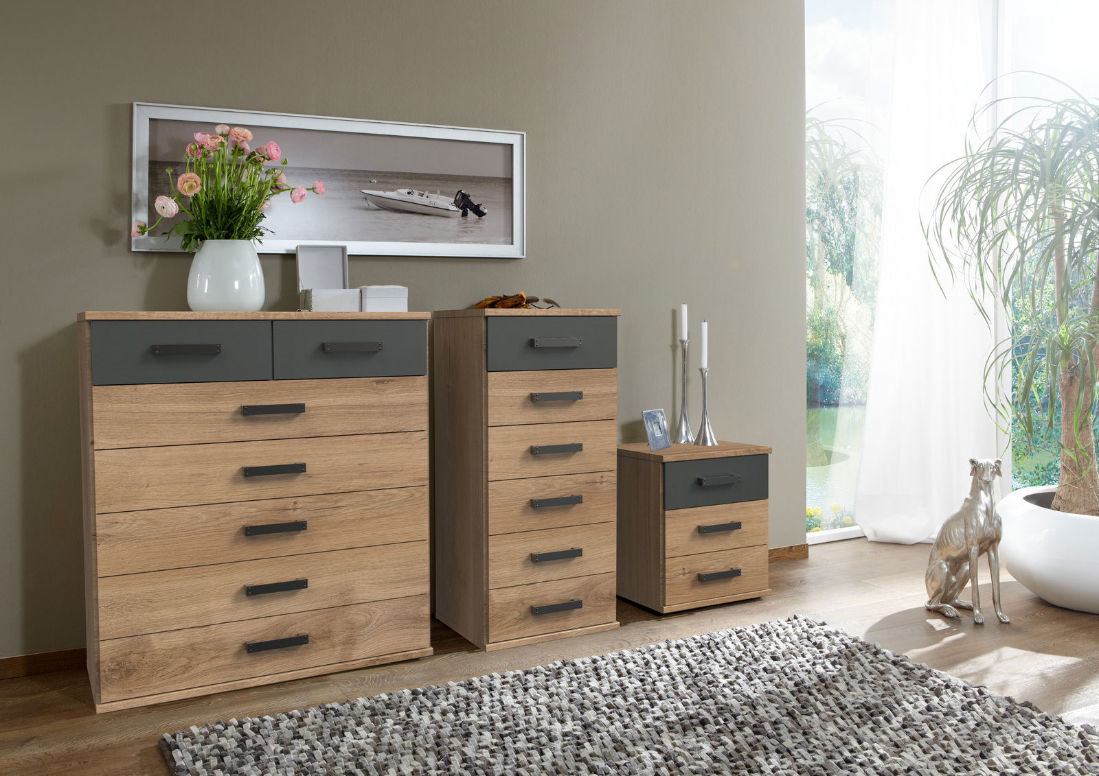 Z2 Kommode CORK Drei Kommoden in verschiedenen Größen aus Holz mit grauen Schubladenfronten, dekoriert mit Blumen und Kerzen, in einem modernen Wohnzimmer mit Blick auf ein Fenster.