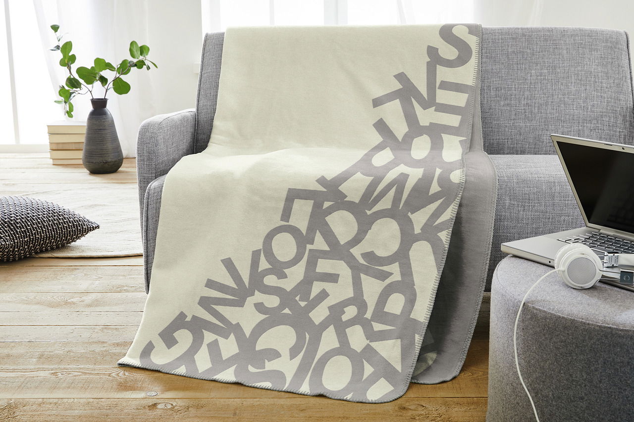 Graue Jacquard-Decke mit Buchstabenmuster auf einem grauen Sofa in einem modernen Wohnzimmer, seitliche Perspektive.