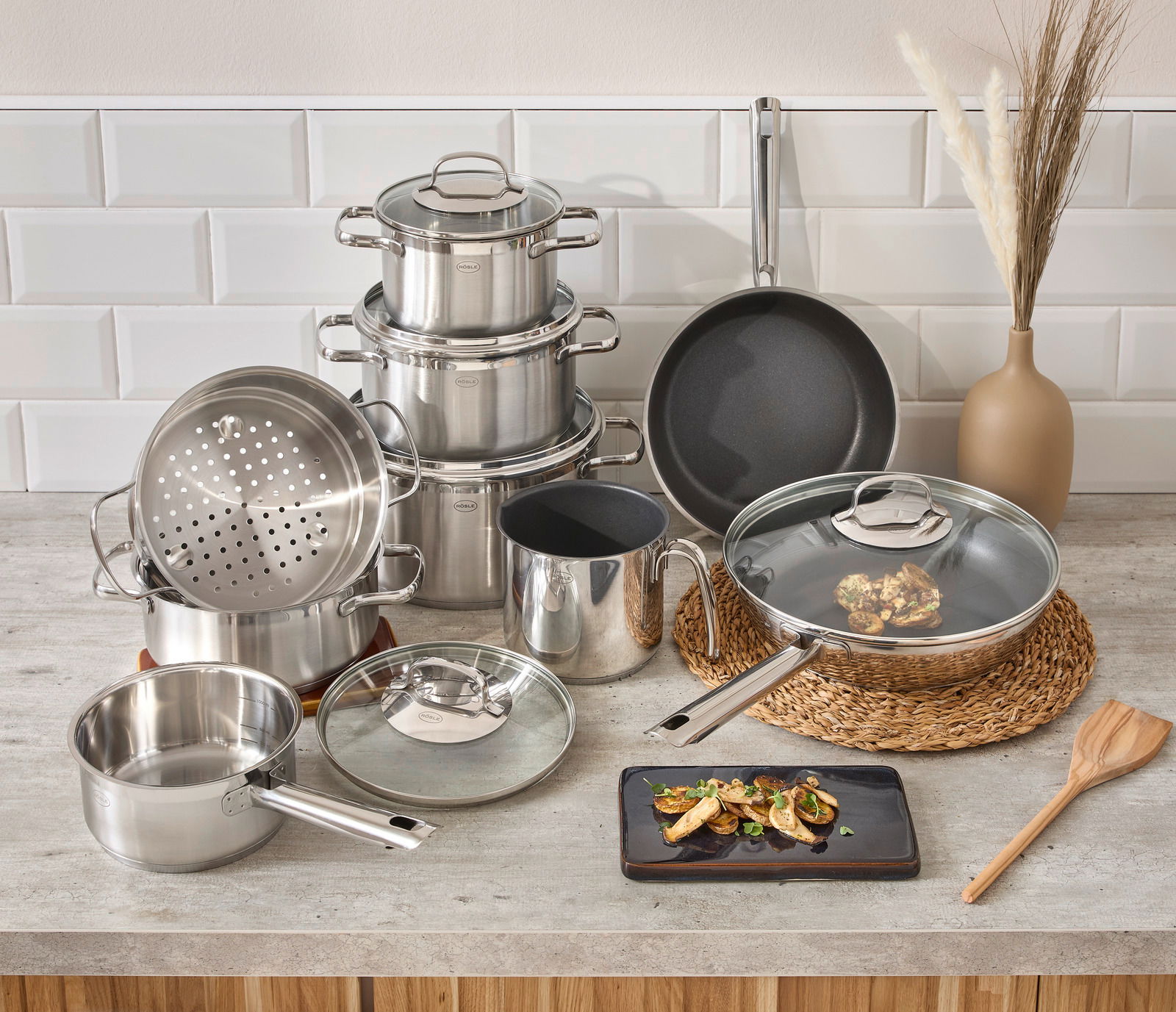 Rösle Topfset 10-tlg. ELEGANCE Topfset Elegance 10-tlg. aus Edelstahl, bestehend aus mehreren Töpfen und Pfannen mit Glasdeckeln, auf einer Küchenarbeitsplatte arrangiert. Perspektive von vorne.