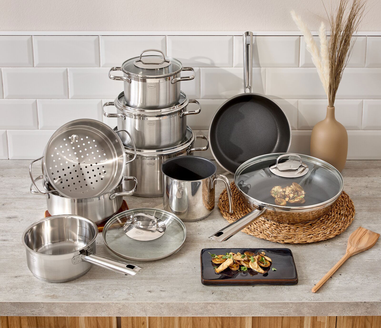 Rösle Topfset 10-tlg. ELEGANCE Topfset Elegance 10-tlg. aus Edelstahl, bestehend aus mehreren Töpfen und Pfannen mit Glasdeckeln, auf einer Küchenarbeitsplatte arrangiert. Perspektive von vorne.