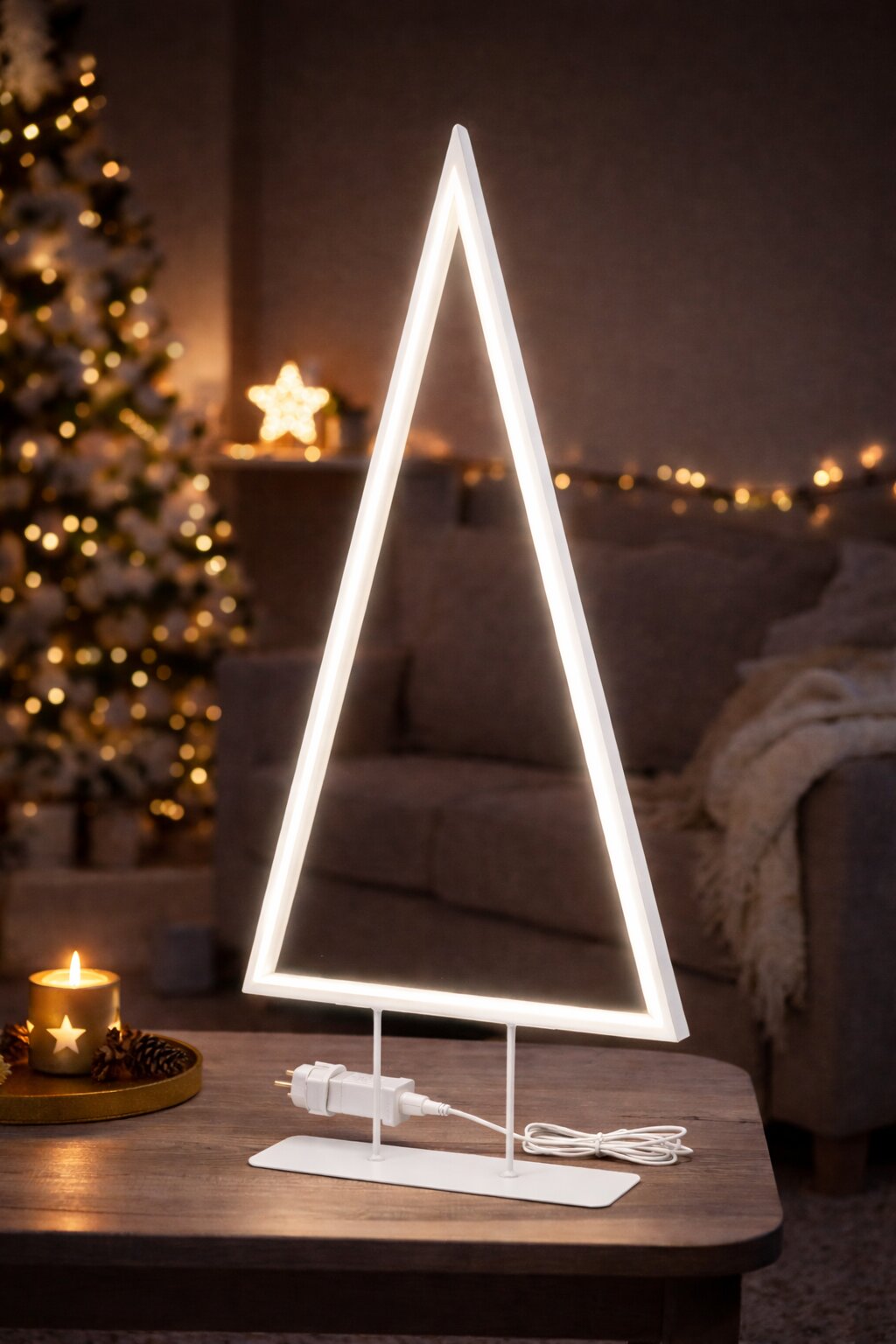 Frontale Ansicht einer leuchtenden, dreieckigen LED-Dekoleuchte in Weiß auf Metallfuß mit Kabel und Netzteil, auf einem Tisch im gemütlich weihnachtlich dekorierten Wohnzimmer.