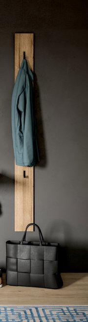 Schmale Garderobenleiste aus hellem Holz mit zwei schwarzen Haken, daran hängt eine blaue Jacke; unten steht eine schwarze geflochtene Tasche vor grauer Wand, Frontansicht.