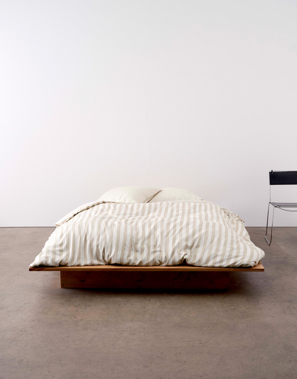 Bed met gestreept beddengoed in beige en wit, van voren gefotografeerd, op een laag houten frame.