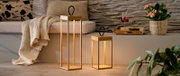 Twee moderne led-lantaarns van goudkleurig metaal met een zwarte handgreep, die een warme gloed verspreiden op de houten vloer voor de bank en de decoratie; perspectief: frontaal, licht zijdelings aanzicht vanaf vloerniveau