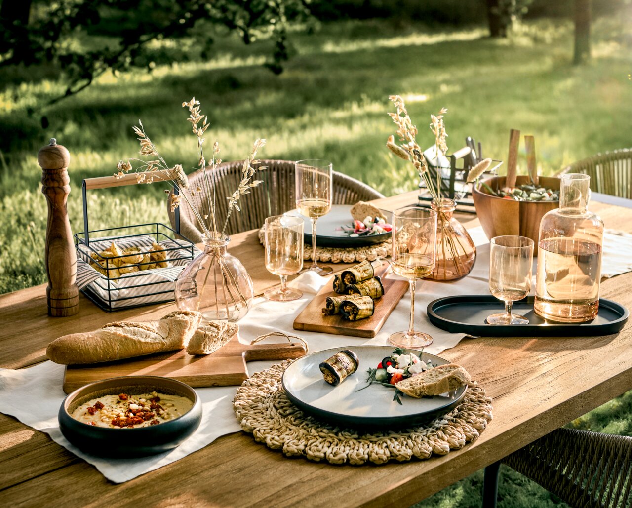 Ein stilvoll gedeckter Tisch im Freien mit einem 16-teiligen Kombiservice, bestehend aus Tellern, Gläsern und Karaffen, umgeben von natürlichem Licht und grüner Landschaft. Perspektive von oben.