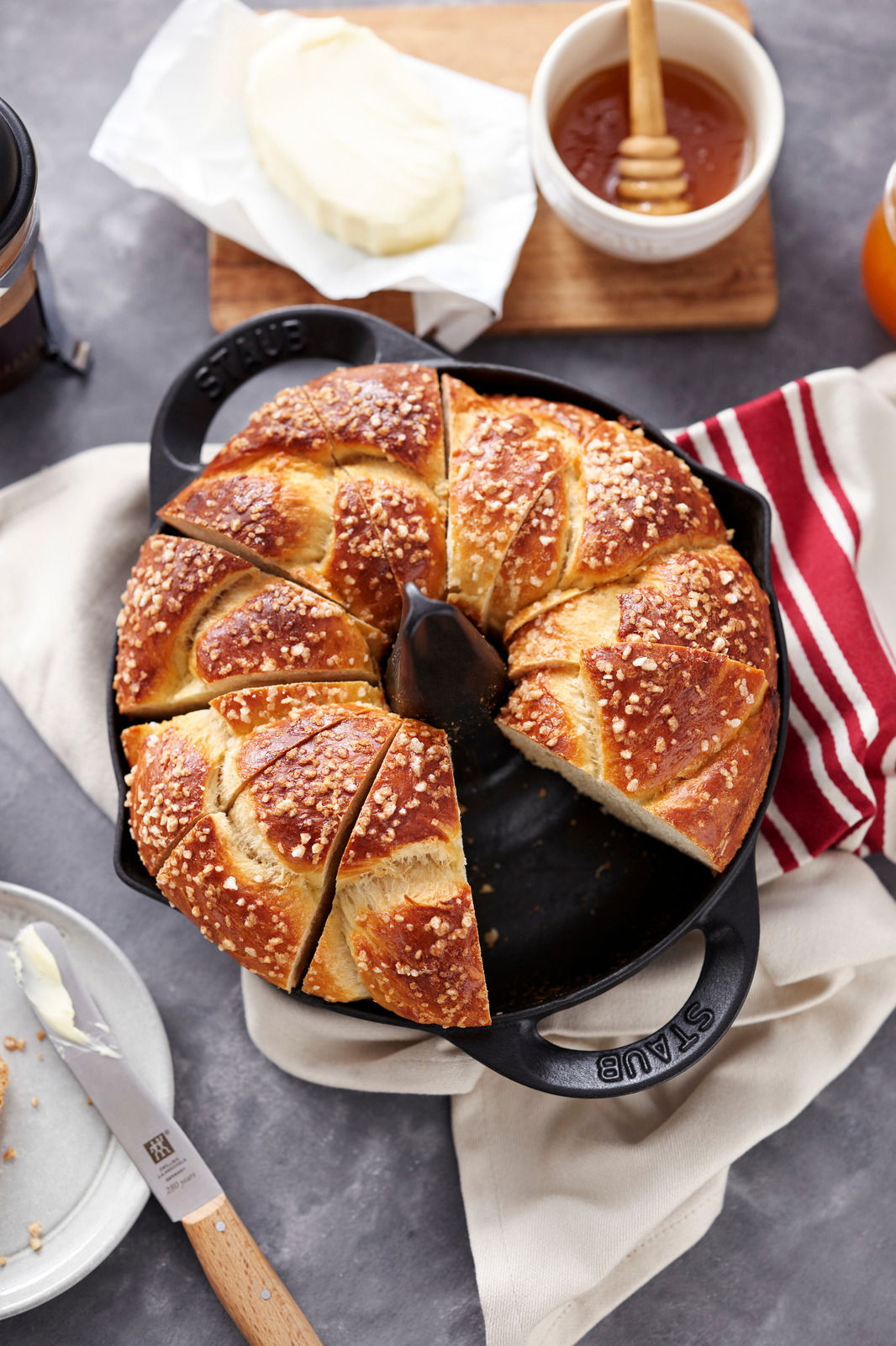 Vogelperspektive auf einen STAUB Hähnchenbräter mit frischem, geschnittenem Gebäck, umgeben von Butter, Honig und einem Messer.
