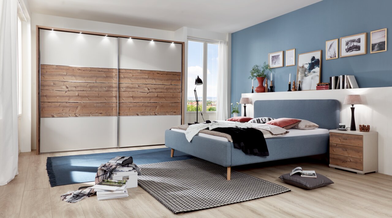 Modernes Schlafzimmer mit einem blauen Polsterbett in Frontalansicht, flankiert von einem Nachttisch aus Holz. Im Hintergrund ein großer Kleiderschrank mit Holz- und Weißakzenten.