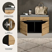 Schuhbank aus hellem Holz mit grauem Sitzkissen, zwei Türen geöffnet und schwarzem Stauraumfach; oben liegen Tasche, Kaffeebecher und Zeitung, Frontansicht in leichter Schrägperspektive.