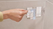 Hand befestigt tesa Lavaa Eckduschkorb M an einer Badezimmerwand mit Klebestreifen, seitliche Perspektive