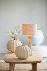 Moderner Lampenfuß aus Holz mit einem schlichten, beigen Lampenschirm, auf einem Holztisch in einem stilvoll dekorierten Raum, seitliche Perspektive.