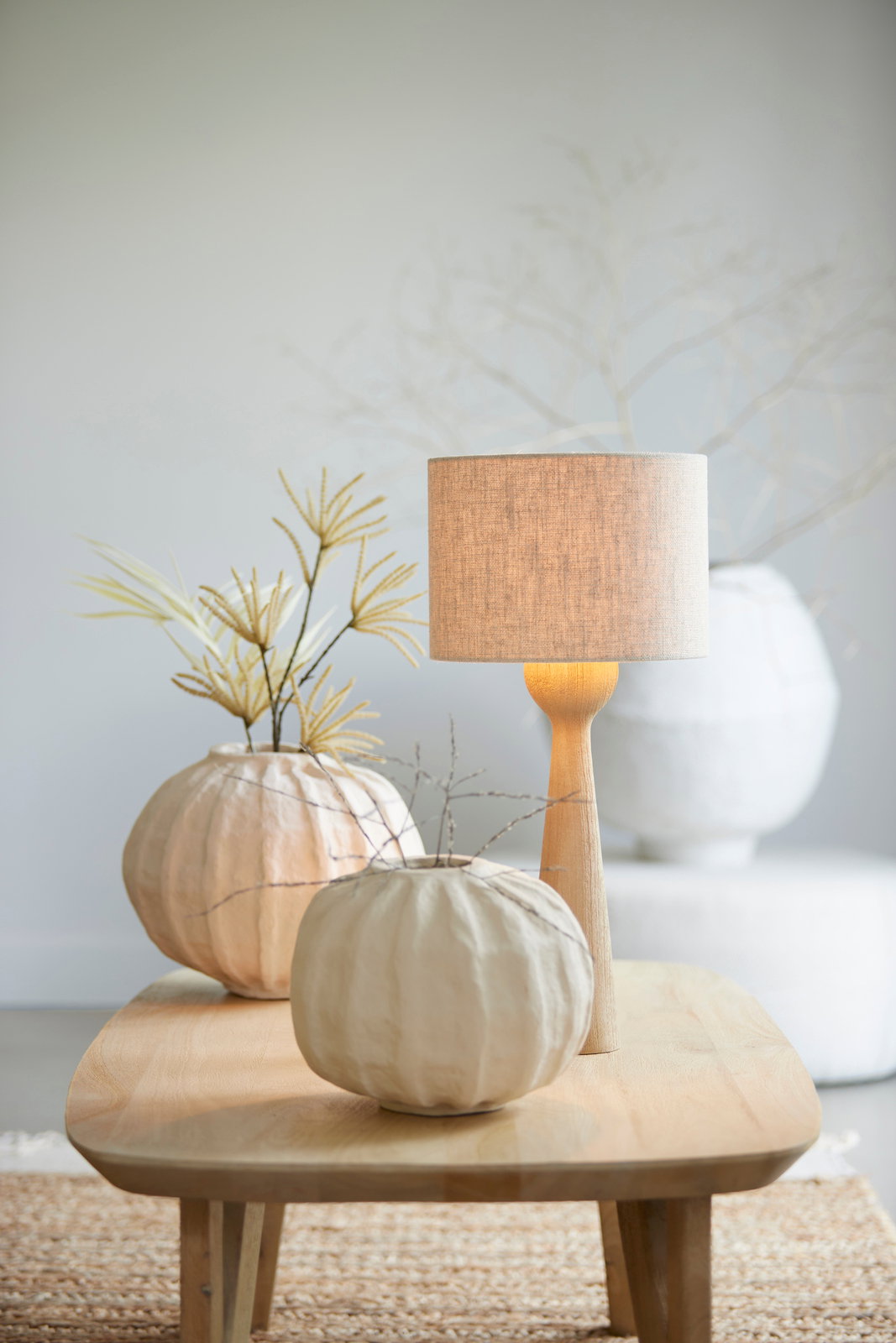 Moderner Lampenfuß aus Holz mit einem schlichten, beigen Lampenschirm, auf einem Holztisch in einem stilvoll dekorierten Raum, seitliche Perspektive.