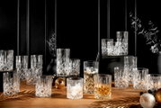 Ein elegantes 18-teiliges Becherset Noblesse aus Kristallglas, präsentiert auf einem Holztisch mit dekorativen Elementen. Die Gläser sind in verschiedenen Größen und mit Getränken gefüllt, in einer stilvollen, festlichen Umgebung arrangiert. Perspektive: