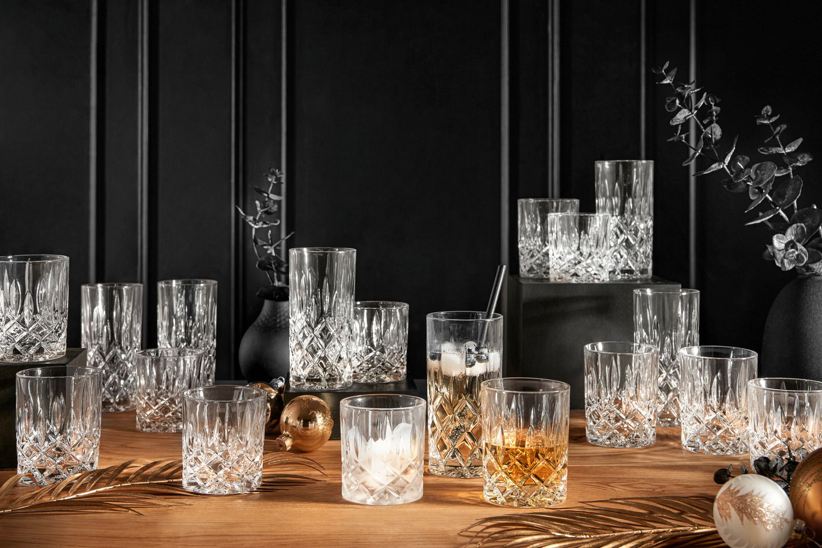 Ein elegantes 18-teiliges Becherset Noblesse aus Kristallglas, präsentiert auf einem Holztisch mit dekorativen Elementen. Die Gläser sind in verschiedenen Größen und mit Getränken gefüllt, in einer stilvollen, festlichen Umgebung arrangiert. Perspektive: 