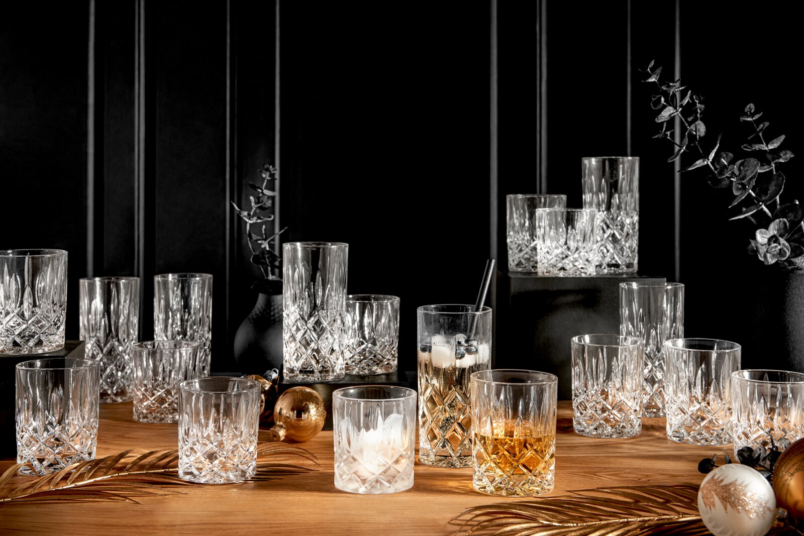 Ein elegantes 18-teiliges Becherset Noblesse aus Kristallglas, präsentiert auf einem Holztisch mit dekorativen Elementen. Die Gläser sind in verschiedenen Größen und mit Getränken gefüllt, in einer stilvollen, festlichen Umgebung arrangiert. Perspektive: 