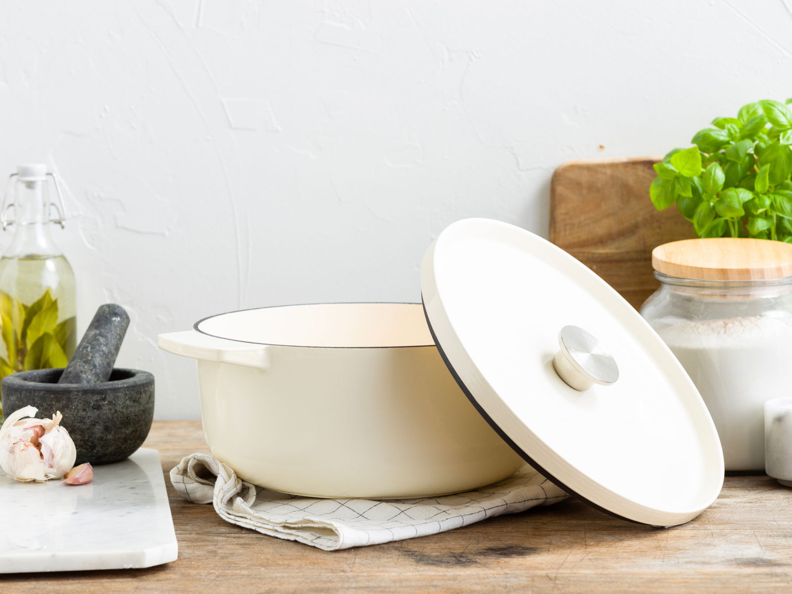 Gusseisen Bräter 26cm in Mandelweiß, seitliche Perspektive, auf einem Küchentisch mit Küchenutensilien im Hintergrund.