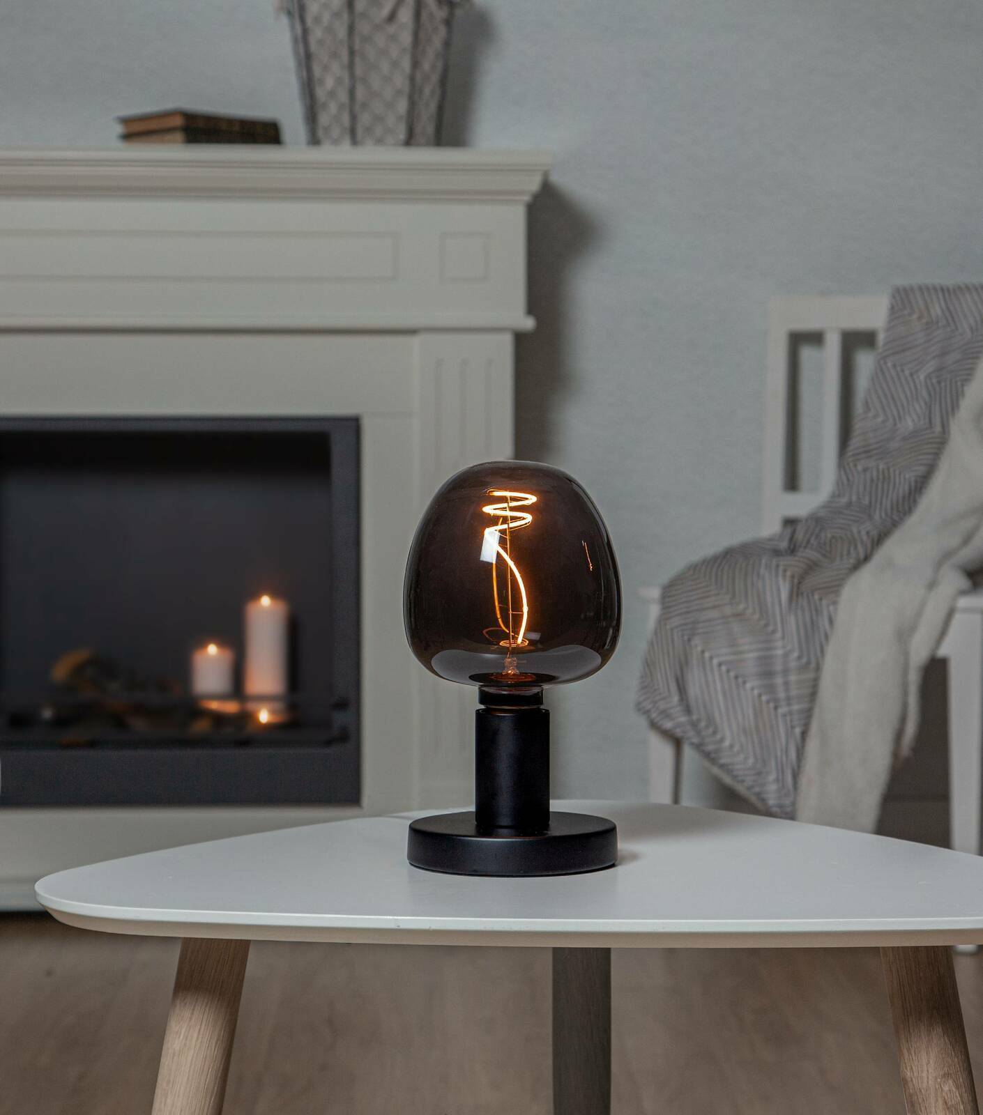 Moderne Tischlampe mit schwarzem Sockel und rauchgrauem Glas, leuchtend auf einem weißen Tisch in einem gemütlichen Wohnzimmer, seitliche Perspektive.