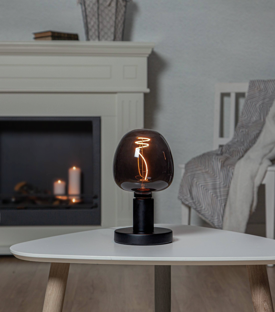 Moderne Tischlampe mit schwarzem Sockel und rauchgrauem Glas, leuchtend auf einem weißen Tisch in einem gemütlichen Wohnzimmer, seitliche Perspektive.