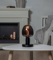 Moderne Tischlampe mit schwarzem Sockel und rauchgrauem Glas, leuchtend auf einem weißen Tisch in einem gemütlichen Wohnzimmer, seitliche Perspektive.