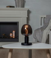 Moderne Tischlampe mit schwarzem Sockel und rauchgrauem Glas, leuchtend auf einem weißen Tisch in einem gemütlichen Wohnzimmer, seitliche Perspektive.