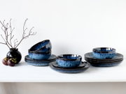 Blau-schwarz glasiertes Keramikgeschirr-Set mit Schalen und Tellern auf weißer Tischplatte, seitliche Frontansicht (leicht von oben) vor hellem Hintergrund; links schwarze Vase mit Zweigen und Früchten.