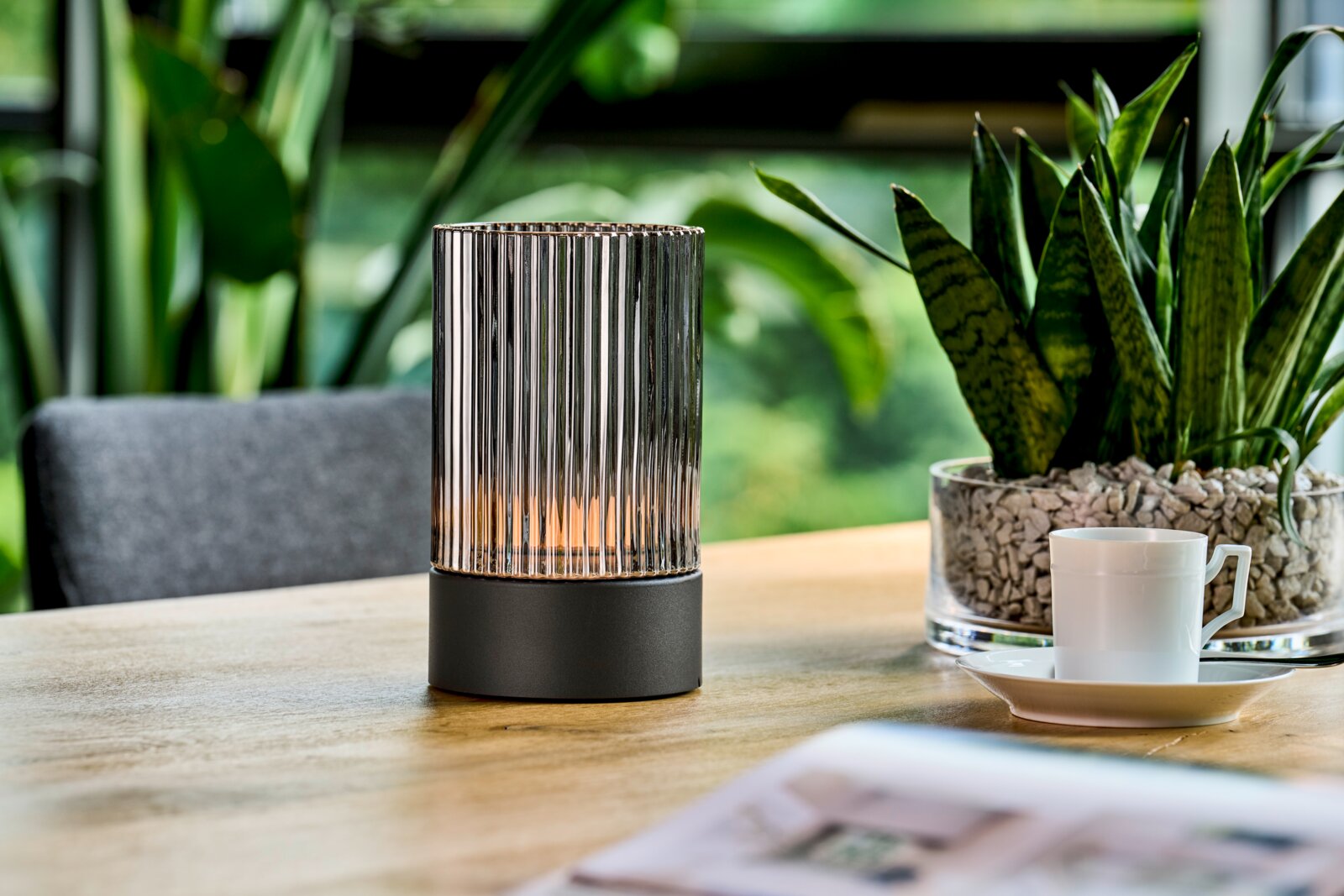 Moderne Akku-Tischleuchte mit geriffeltem Glasdesign auf einem Holztisch, umgeben von Pflanzen, aus seitlicher Perspektive.