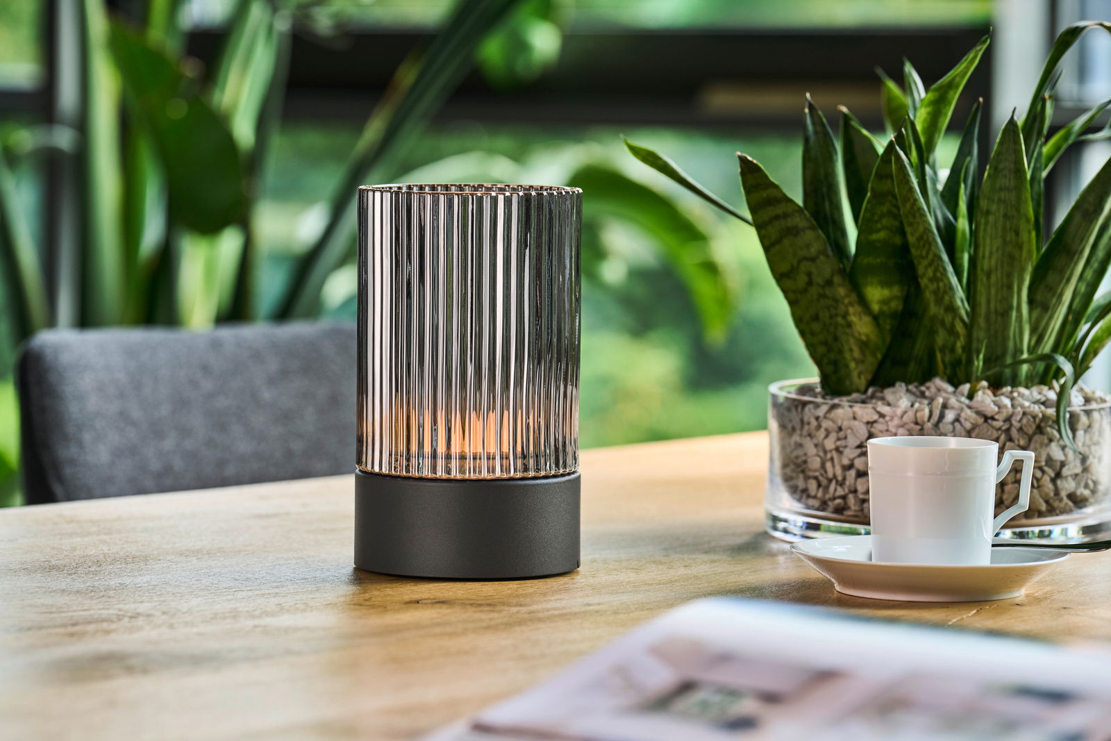 Moderne Akku-Tischleuchte mit geriffeltem Glasdesign auf einem Holztisch, umgeben von Pflanzen, aus seitlicher Perspektive.