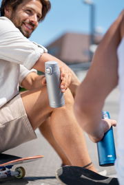 Zwei Personen sitzen im Freien, eine hält einen silbernen Thermobecher in der Hand, während die andere einen blauen Becher hält. Perspektive von der Seite.