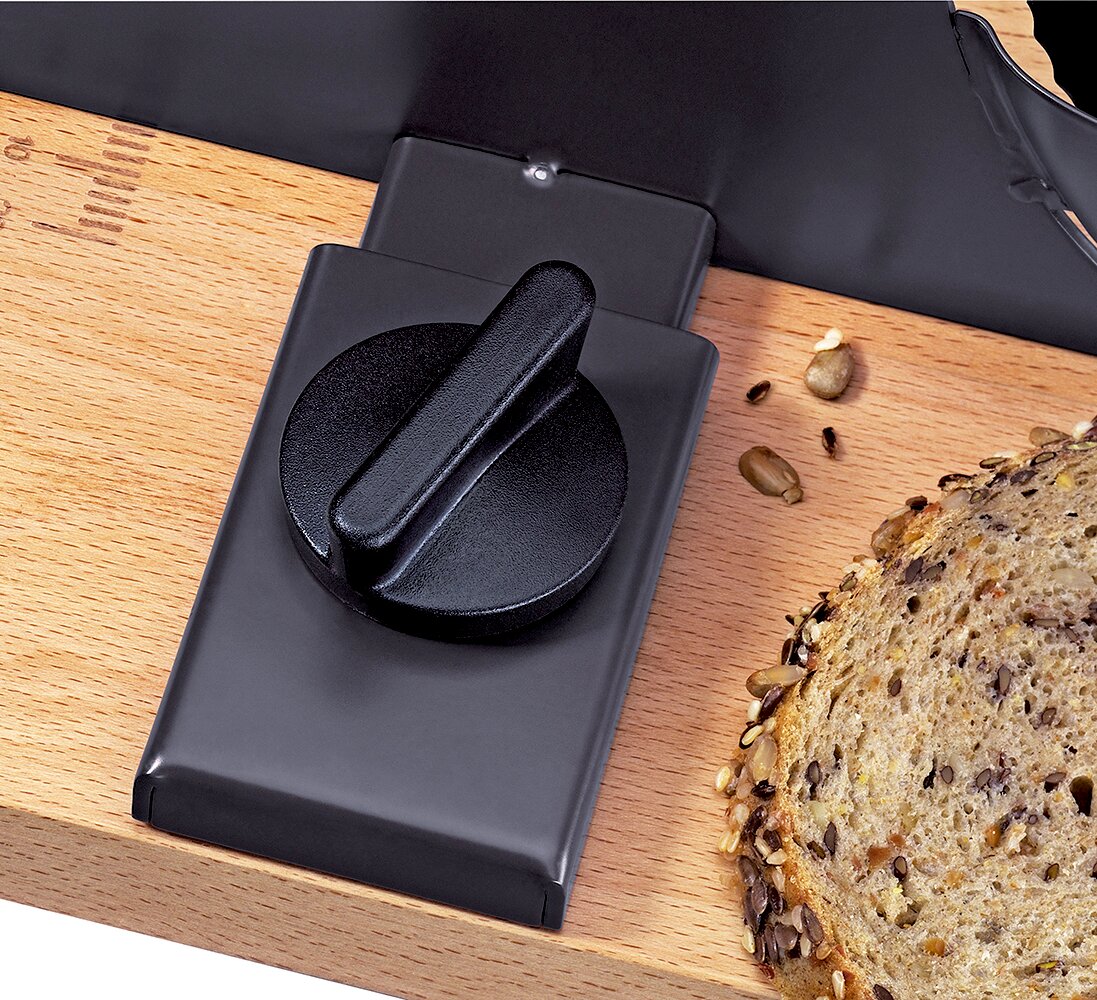Nahaufnahme aus schräger Draufsicht: schwarzer Drehknopf einer Brotschneidemaschine auf einem Holzbrett neben einer Scheibe Körnerbrot und verstreuten Samen.