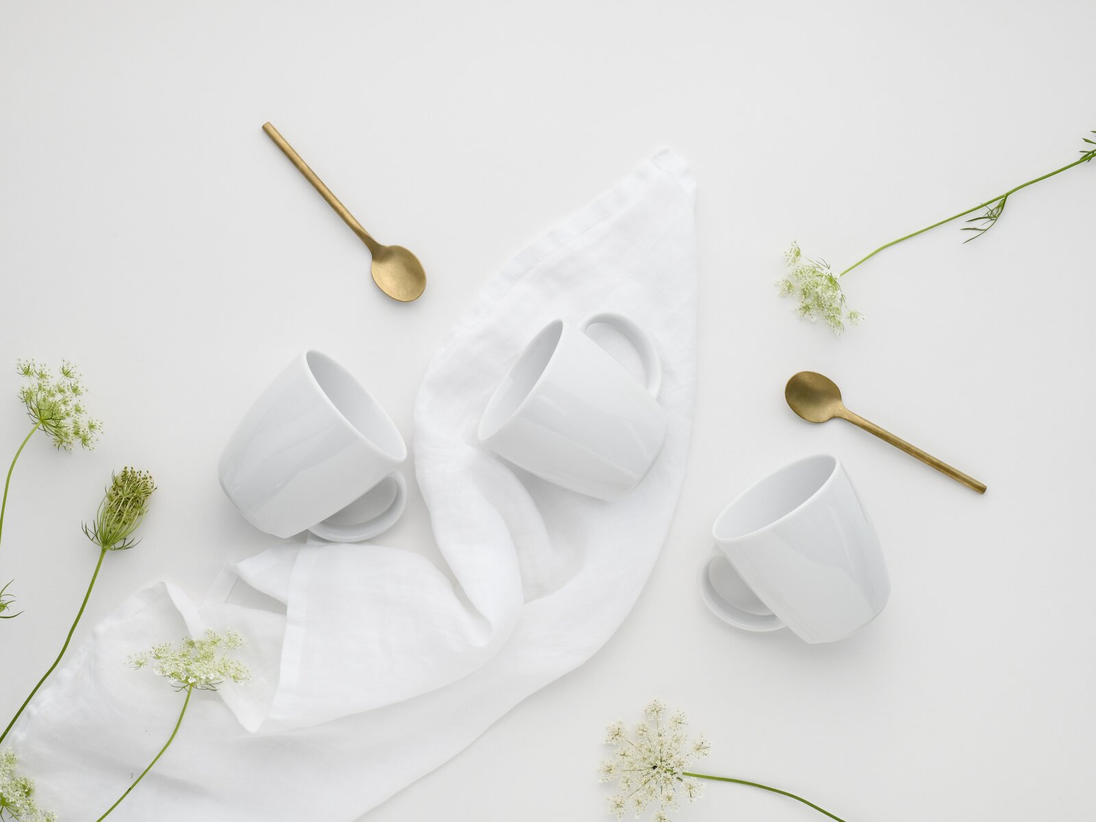 Minimalistisches Flatlay aus der Draufsicht: drei weiße Kaffeetassen mit Untertassen auf weißem Tuch, dazu zwei goldene Löffel und zarte weiße Blüten auf hellem Hintergrund.