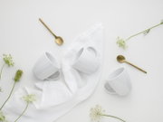 Minimalistisches Flatlay aus der Draufsicht: drei weiße Kaffeetassen mit Untertassen auf weißem Tuch, dazu zwei goldene Löffel und zarte weiße Blüten auf hellem Hintergrund.