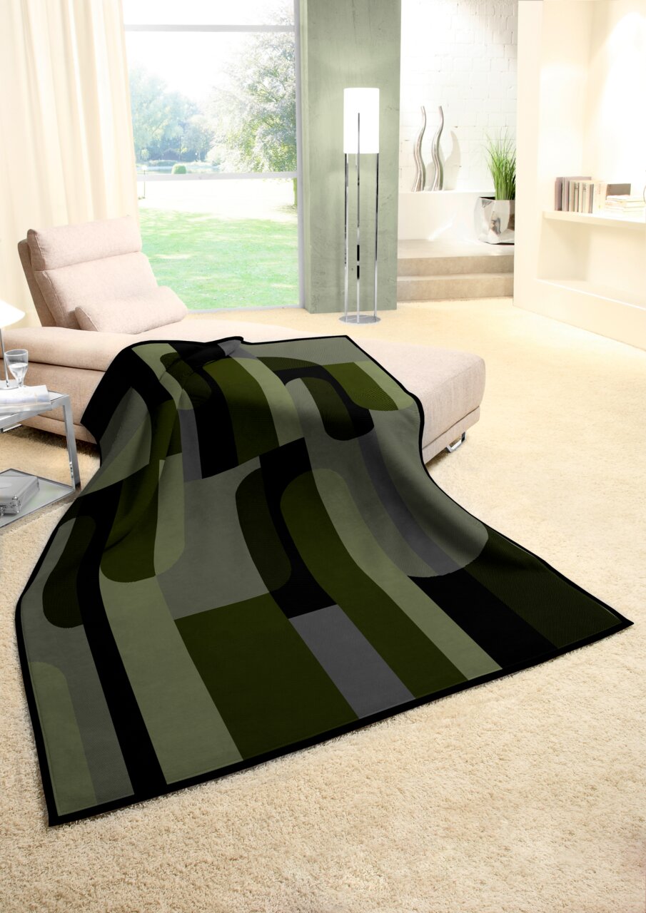 Moderne Wohndecke in Grün- und Grautönen mit geometrischem Muster, drapiert über einem Sofa in einem hellen Wohnzimmer mit Blick auf den Garten.