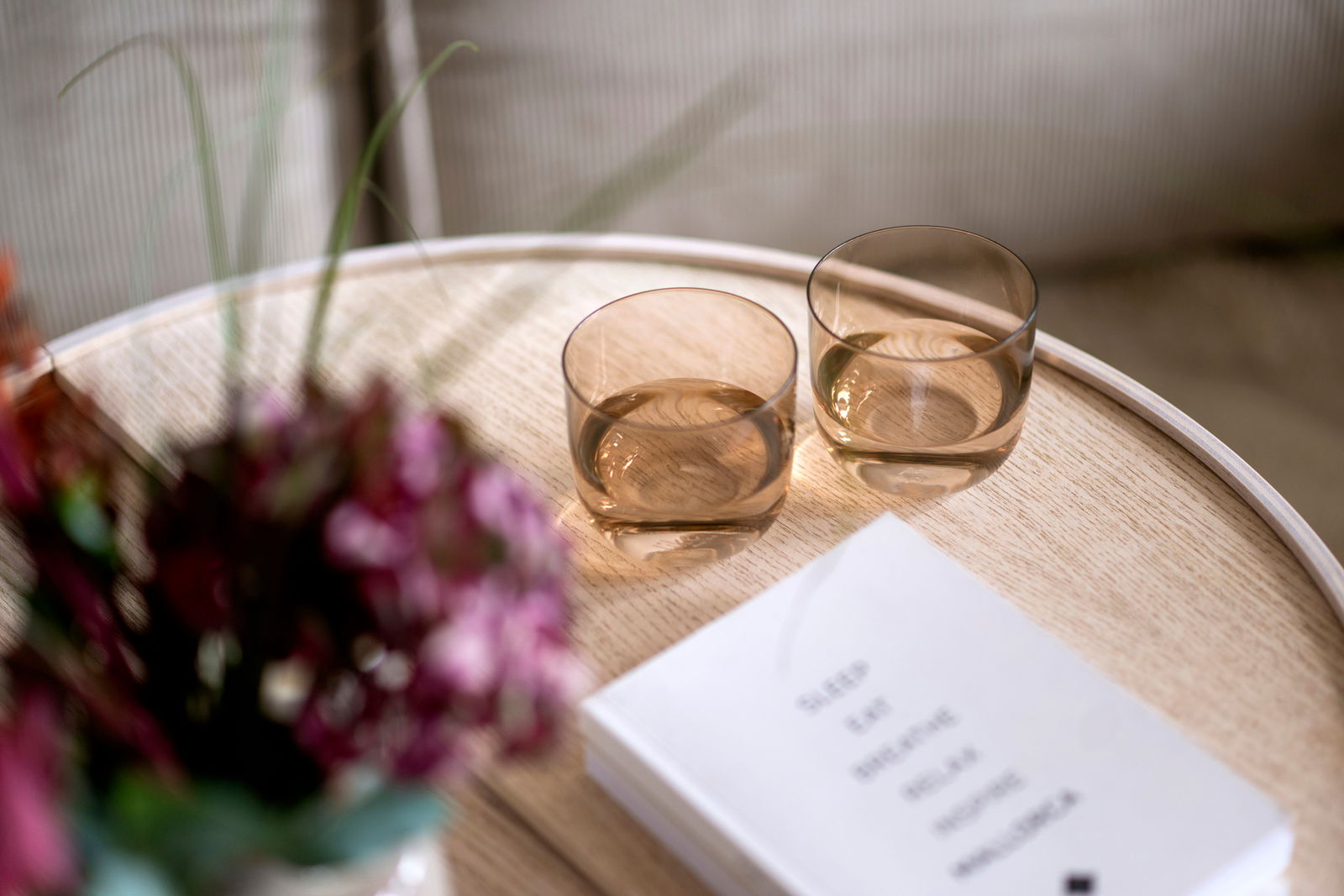 Zwei Wassergläser auf einem runden Holztisch, umgeben von einem Blumenstrauß und einem Buch, aus der Vogelperspektive fotografiert.