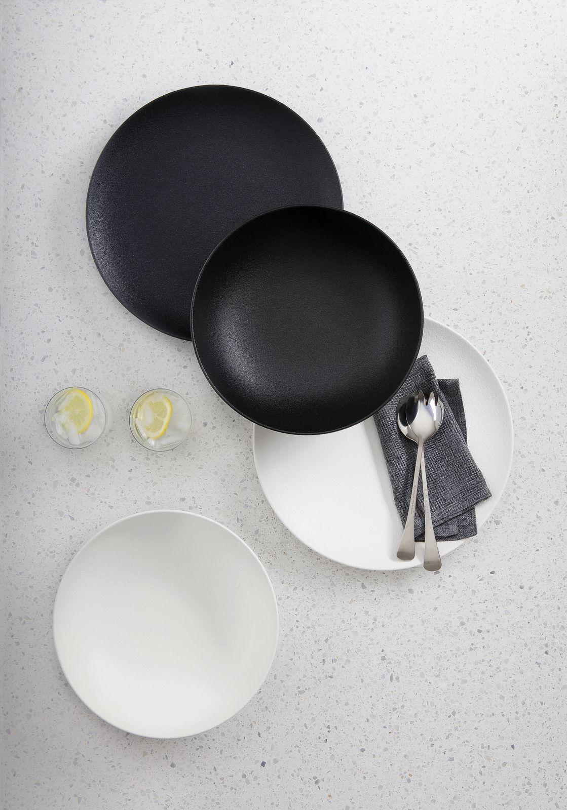 Vogelperspektive auf ein 8-teiliges Dinner-Set in Schwarz und Weiß, bestehend aus Tellern und Schalen, mit Besteck und Servietten auf einem hellen Untergrund.