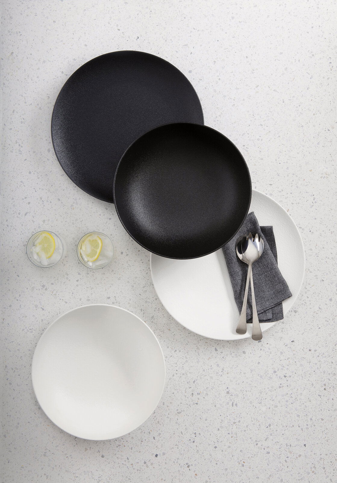 Vogelperspektive auf ein 8-teiliges Dinner-Set in Schwarz und Weiß, bestehend aus Tellern und Schalen, mit Besteck und Servietten auf einem hellen Untergrund.