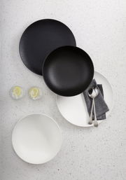 Vogelperspektive auf ein 8-teiliges Dinner-Set in Schwarz und Weiß, bestehend aus Tellern und Schalen, mit Besteck und Servietten auf einem hellen Untergrund.