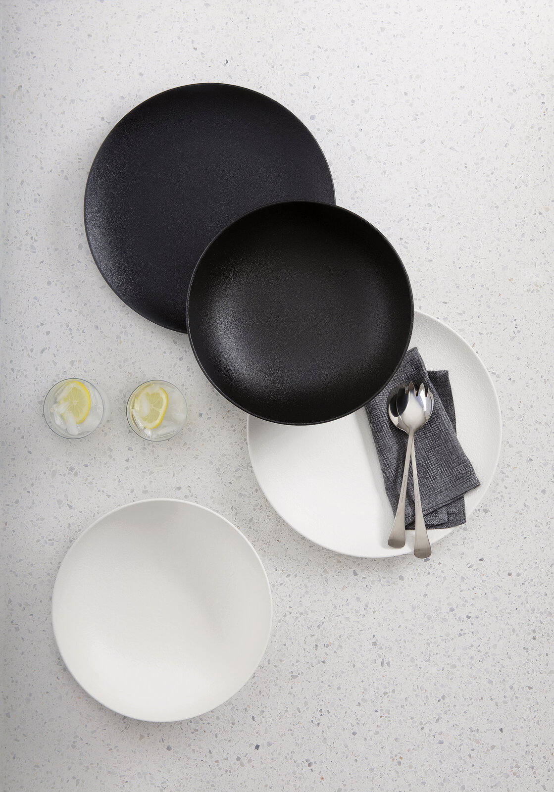 Vogelperspektive auf ein 8-teiliges Dinner-Set in Schwarz und Weiß, bestehend aus Tellern und Schalen, mit Besteck und Servietten auf einem hellen Untergrund.