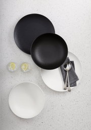 Vogelperspektive auf ein 8-teiliges Dinner-Set in Schwarz und Weiß, bestehend aus Tellern und Schalen, mit Besteck und Servietten auf einem hellen Untergrund.